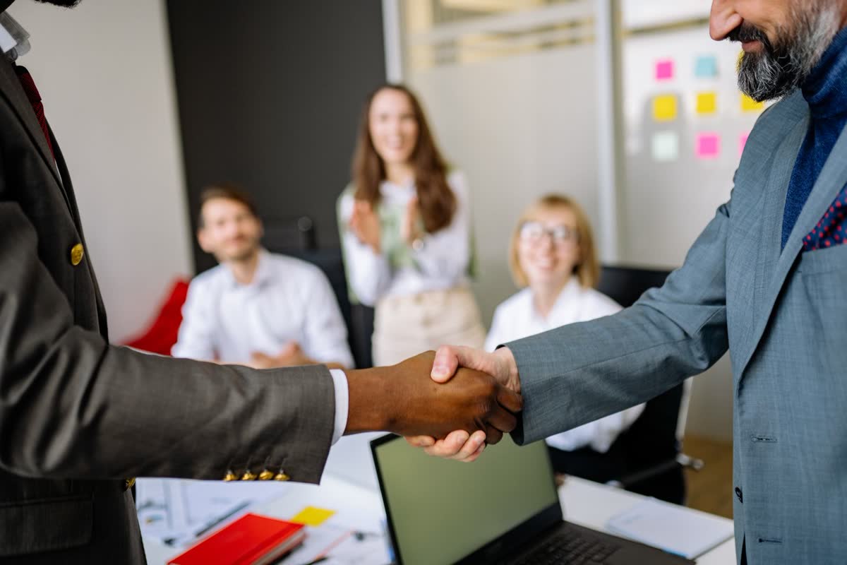 Two professionals shaking hands across a desk in a bright office while colleagues look on