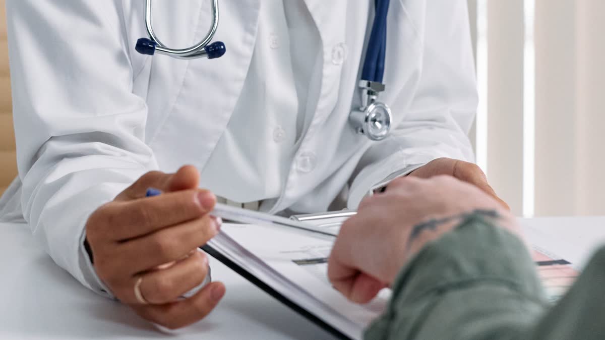 A doctor in a white coat with a stethoscope reviewing a clipboard with a patient across a desk