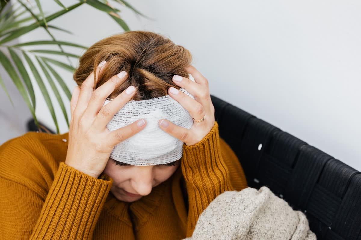 Woman with a bandage wrapped around her head and both hands on her head, sitting indoors