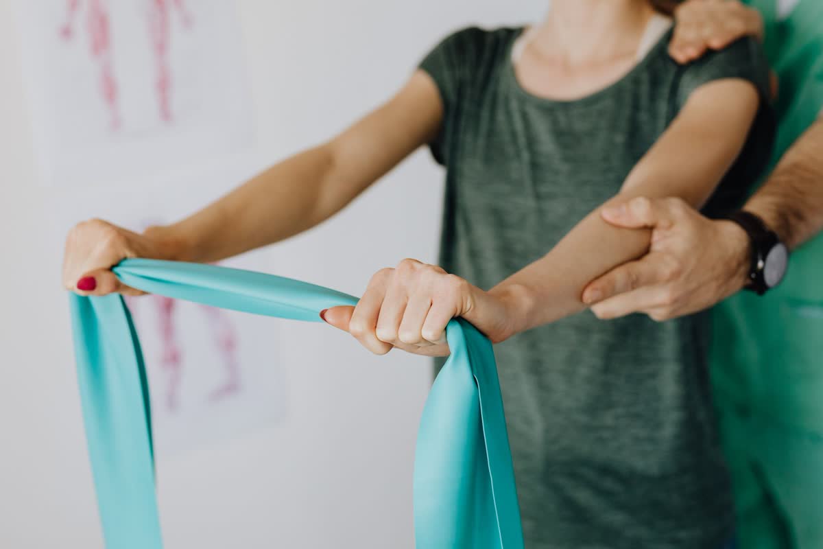 A patient pulling a teal resistance band while a clinician supports her shoulder during a rehab session