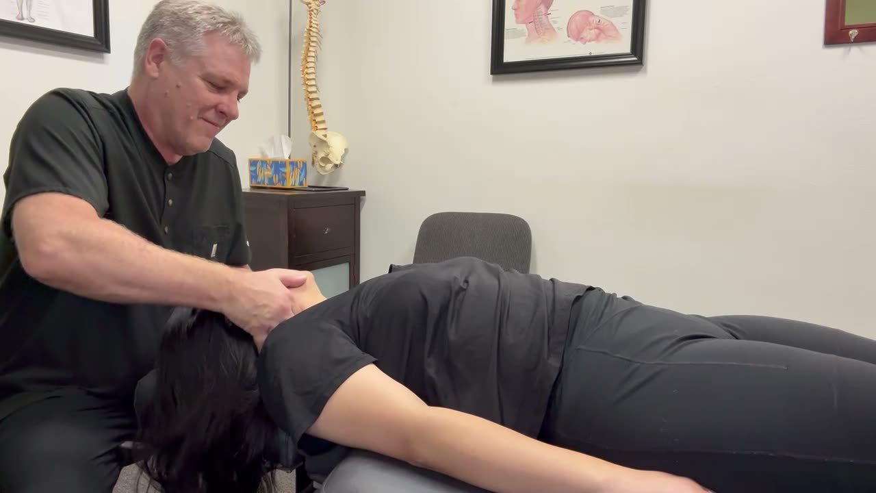 A chiropractor in dark scrubs gently positioning a patient's neck as she lies face down on a treatment table, with an anatomical spine model and wall charts behind them